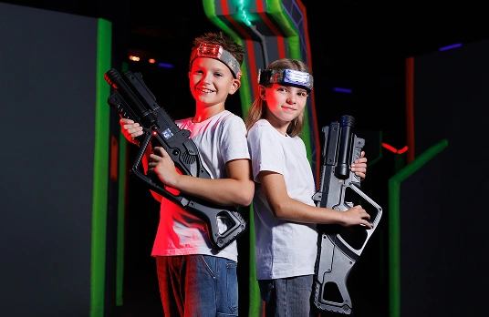 Op de foto staan twee lachende kinderen — een jongen en een meisje — die lasertag spelen. Ze staan rug aan rug tegen de achtergrond van een fel verlichte neonarena.